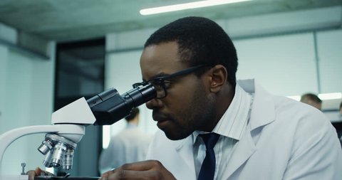 Close up of the young attractive African American man, medical researcher, looking in the microscope while doing some analysis and typing them in the laptop computer at the laboratory.