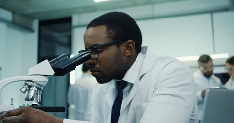 African american young male scientist doing some research in the laboratory while looking in the microscope and working at the laptop computer. Close up.  2019-ncov, coronavirus, virus 