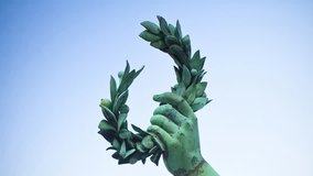 Hand of an ancient bronze statue holds a laurel wreath against a sky background