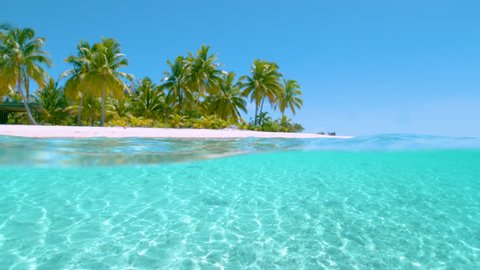 SLOW MOTION HALF IN HALF OUT: Stunning shot of the calm ocean surface and the idyllic tropical island sandy beach with lush palm trees towering into the clear blue sky. Picturesque One Foot Island.