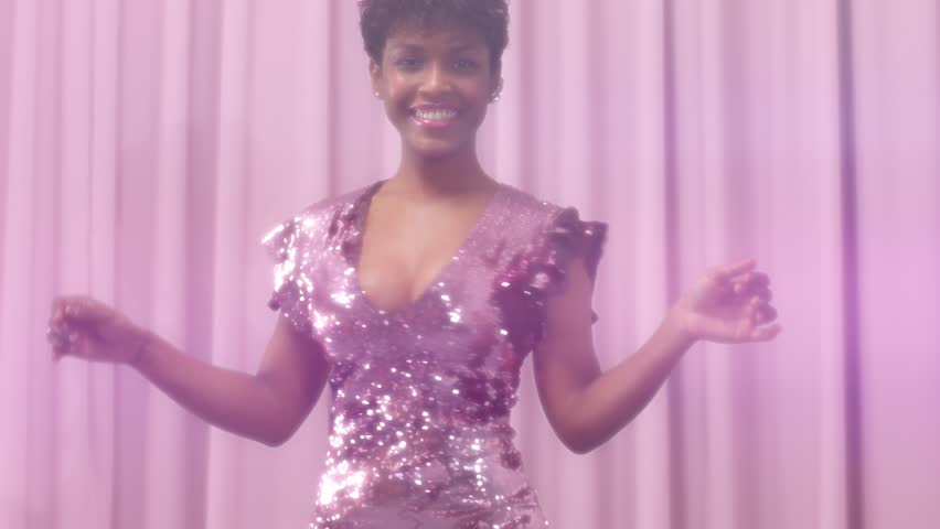 black mixed race woman with short haircut and curly natural hair wears sequin sparkly dress in pink. Dancing womn and sparkly sequin dress makes lens flair