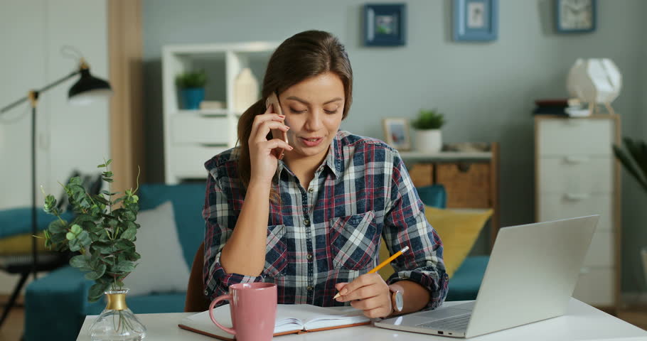Caucasian pretty young woman smiling and talking on the phone at the table with a laptop computer in the cozy room.