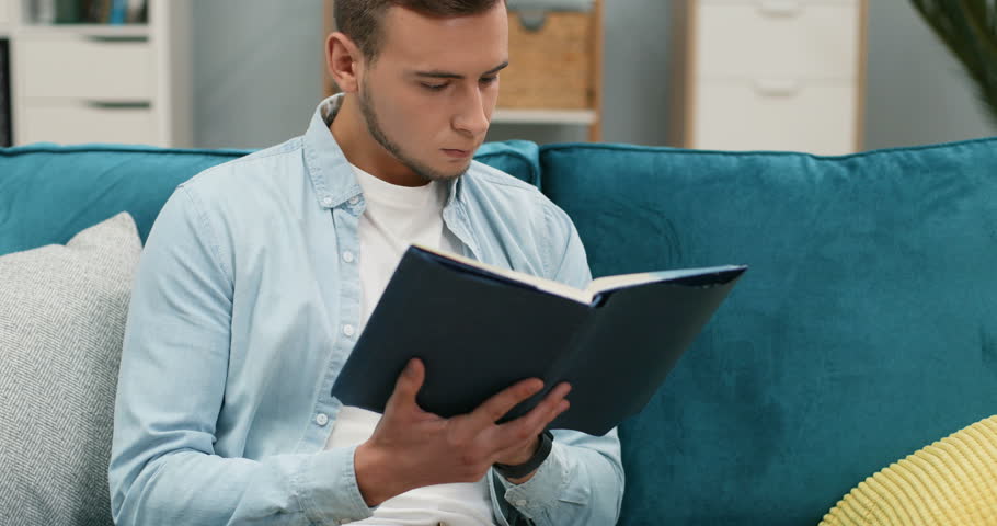 Close up of the Caucasian guy student reading a book and studing on the couch at home.