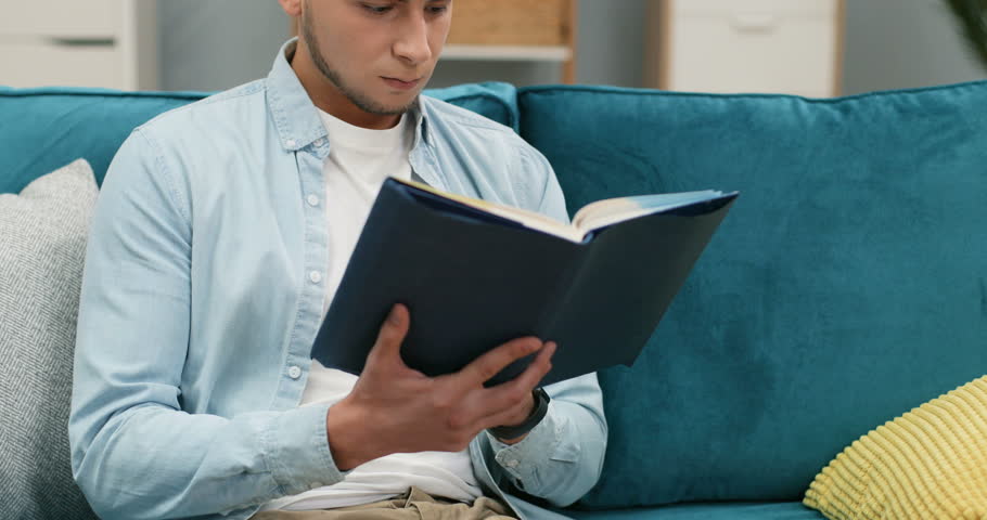 Portrait of the Caucasian young guy student reading a textbook and studing on the couch at home, then smiling to the camera. Close up.
