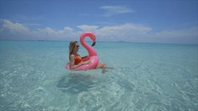 Young woman on tropical beach having fun splashing water with feet. Girl in inflatable flamingo on holidays 