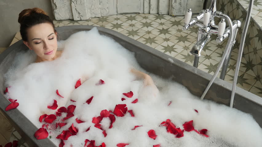 girl lies in a large stone bath in foam with red rose petals next to a large window