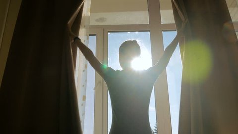 Woman silhouette opening curtains and looking out of window in hotel room at morning. Sun lens flare