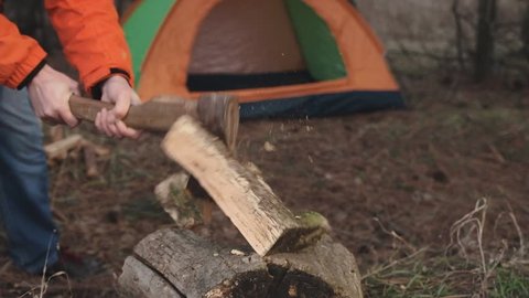 A traveler in a bright jacket and winter hat cuts firewood for making a campfire on the background of a tourist tent set up for an overnight stay in the forest. Slow motion