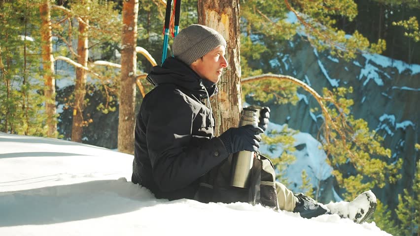 Young man drinking tea or coffee in snowy winter forest. Mountain camping concept. Traveler man hiking in forest in winter time using thermos holding cup of tea.