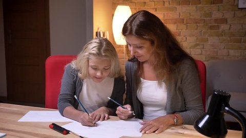 Closeup portrait of young mother and her little pretty daughter playing a tic tac toe game on the paper together at cozy home indoors