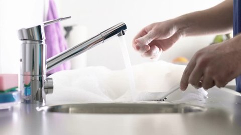 Man washing dishes in the kitchen sink. Person doing domestic household chores. Turn off the water from tap and start to scrub white plate with brush. Soap foam and bubbles.