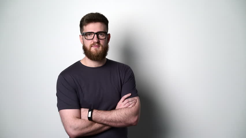 Half length portrait of young bearded hipster man standing with folded hands by the white wall wearing eyeglasses, smiling at camera
