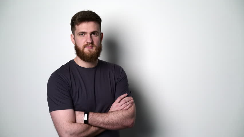 Half length portrait of young bearded hipster man standing with folded hands by the white wall smiling at camera