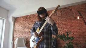 Portrait of young redhead man musician playing electric guitar in VR glasses at home