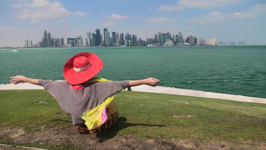 Caucasian woman backside with open arms sitting on green lawn of Doha West Bay in Qatar. Blonde tourist enjoys Doha Downtown from West Mound-Skyline viewpoint. Persian Gulf, Arabian Peninsula.