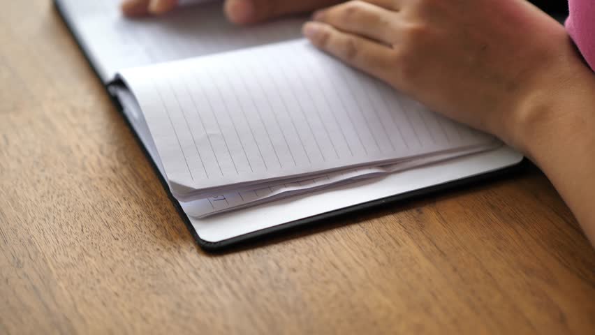 A closeup view of a black mixed race African American woman wearing a pink shirt sitting at a wood dining table and writing a note in her paper notepad with a pen with her hands and chest in frame.