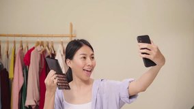 Happy beautiful young Asian woman using smartphone selfie live streaming online to social network. Beauty blogger present beauty cosmetics sitting in front tablet camera for recording video.