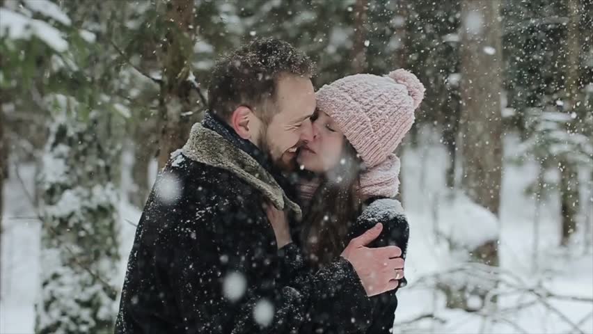 Young beautiful caucasian couple kissing under a snow in a winter forest. Fashionable millennials having fun toghether. Candid footage of young couple in forest. Slow motion.