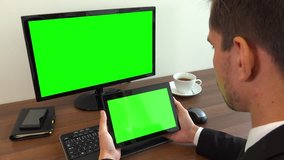 An office worker sits at a desk and looks at a computer and a tablet with green screens - closeup
