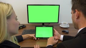 Two office workers, man and woman, sit at a desk and slide at a computer and a tablet with green screens - closeup