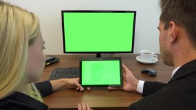 Two office workers, man and woman, sit at a desk and look at a computer and a tablet with green screens - closeup