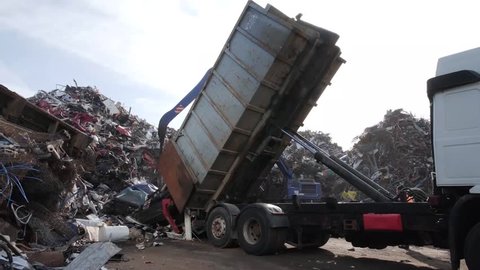 Tipper truck brings car wrecks to a waste landfill site while a crane grabber sorting wrecks in the sunshine