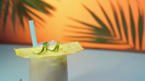 close up view of pina colada cocktail in wet glass with decoration rotating on orange background with green palm leaves