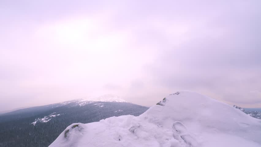 A young man, a tourist with a backpack, stands on the edge of the mountain next to the cliff, arms outstretched to the side, and admires the view of the top of the mountain. Winter season.