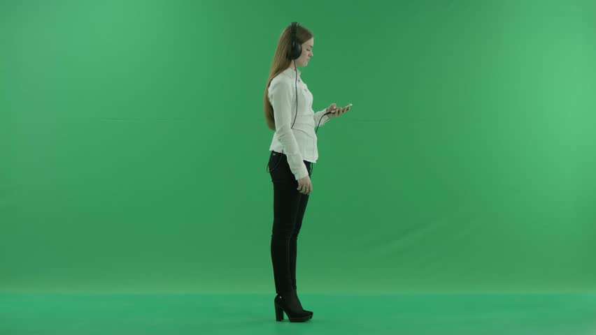 Happy businesswoman listening to music in the headphones and dancing on the spot. She wears formal dress: white shirt and blak trousers, standing in the centre the greenscreen