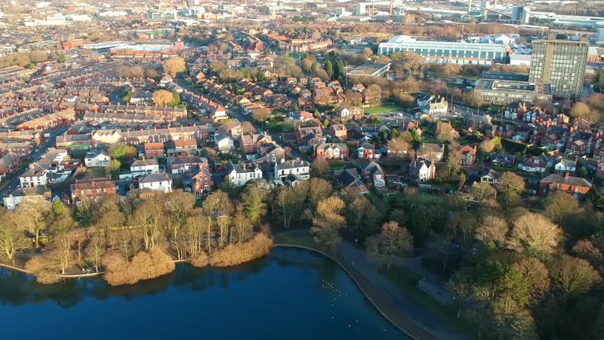 Panning aerial view over public park lake & industrial town suburbs in UK.