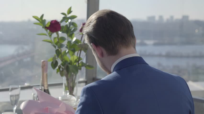 Handsome guy in a stylish jacket sitting with his back to the camera at a table in a cafe or restaurant. The man turns around and meets the girl with a joyful smile on his face.