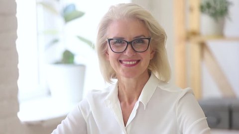 Confident mature middle aged businesswoman in glasses with beautiful face looking at camera posing in office, happy smiling old senior female professional lawyer coach teacher business video portrait