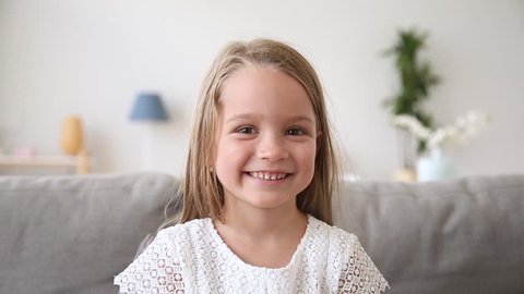 Funny little girl smiling looking at camera at home, cute kid talking to webcam making online video call or recording vlog having fun, preschool child with pretty face waving hand sitting on sofa