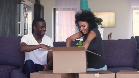 Happy black couple open unpacking cardboard box parcel discussing interior design decor after relocation renovation, african family sitting on couch in living room settle in on moving day in new home
