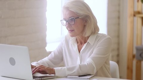 Old middle aged businesswoman working with laptop and papers, busy senior mature woman paying bills online banking managing finances checking budget doing paperwork using computer sitting at desk