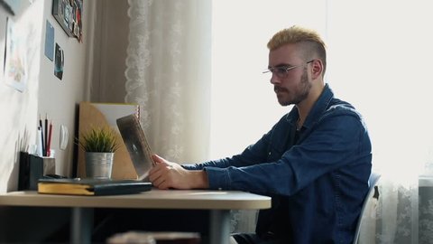 Portrait of handsome young man in glasses with yellow hair shows thumb at the camera. Home Freelancer working at home. Using a notebook. Stylish Successful man working at desk.