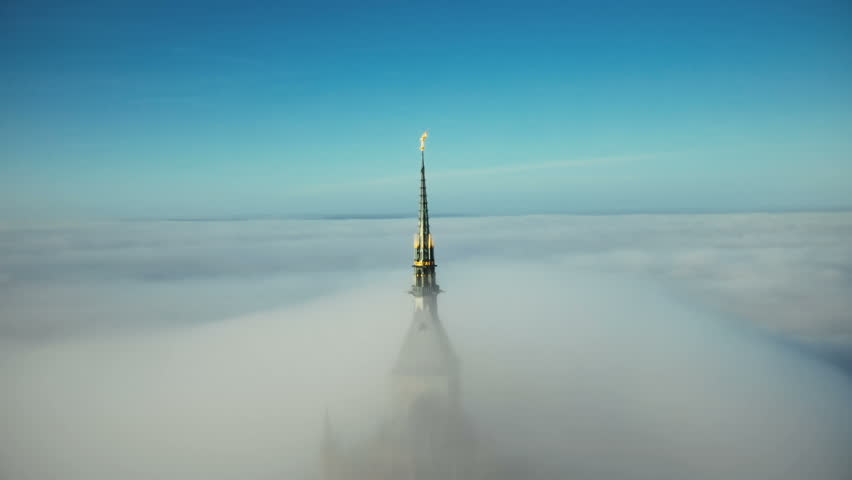 Beautiful aerial panning shot of glorious golden spire at Mont Saint Michel castle covered by epic sunrise fog clouds.