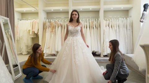 A future bride checks herself out in mirror. She is anxious but very happy and looks forward to her wedding day which is best day in her life. Bridesmaid and shop assistant fix bottom of bride's dress