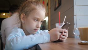 Cute little girl using on the smart phone at the cafe. Happy child having fun and relaxing after school. Natural indoor lifestyle.