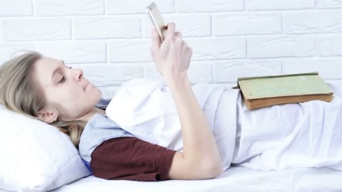 Beautiful young girl in bed makes choice between book and phone. The choice between study and phone. Phone versus book.