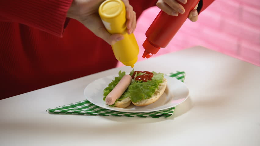 Hungry female pouring ketchup and mustard on hotdog sausage, food flavoring