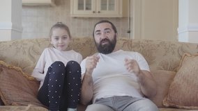 Bearded man sitting on the sofa watching TV with his little daughter supporting football team close up. Girl jumps up when player scored a goal. Concept of happy family. Small girl have fun with dad
