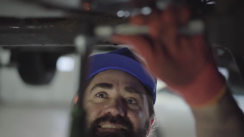 Professional bearded car mechanic screwing details of car with special tool on lifted automobile at repair service station close up. Skillful man in uniform fixing car. Car service, repair maintenance Royalty-Free Stock Footage #1025618717