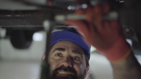 Professional bearded car mechanic screwing details of car with special tool on lifted automobile at repair service station close up. Skillful man in uniform fixing car. Car service, repair maintenance