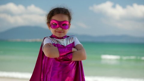 Beautiful Little Girl in the Superhero Costume, Dressed in a Pink Cloak and the Mask of the Hero. Plays on the Background Sea and Blue Sky and Clouds, Sends a Fist Forward. Concept of a Happy