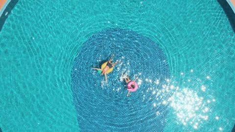 Drone view of young couple floating on swimming pool with inflatable mattresses enjoying their vacations in tropical paradise. Two people relaxing on holidays having fun in summer days sunbathing.