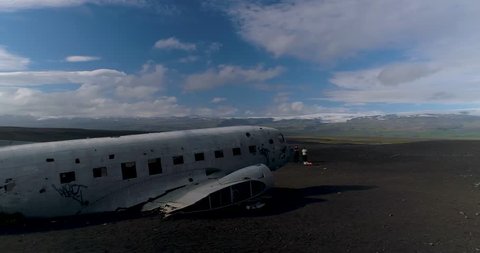 4K low close drone fly by of plane wreck on black sand beach in Iceland. Quick orbit back to nose of plane. Green mountains in background. Cloudy blue sky. Unidentifiable tourists standing.