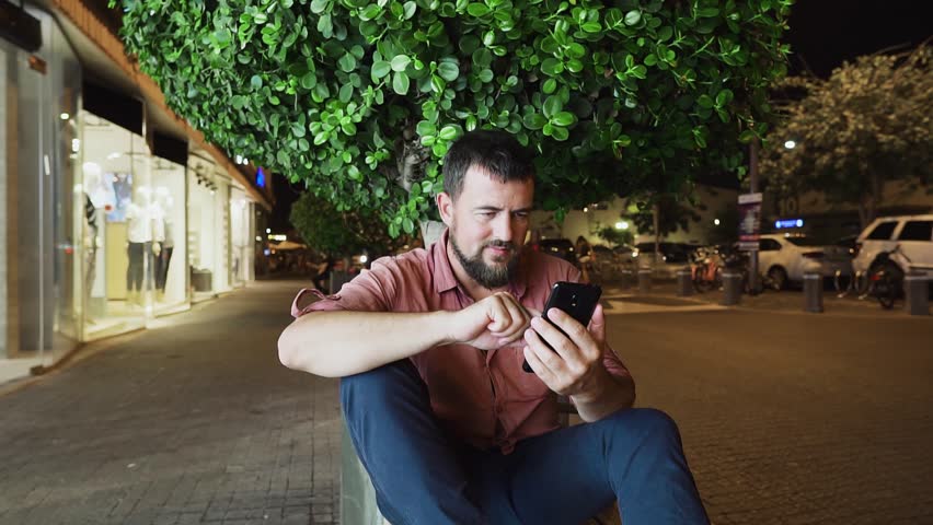 Attractive stylish young man scrolls his cellphone and smiles. Handsome young man sitting on the street and using cell phone at night.