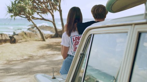 Young couple hanging out on beach looking at ocean beside car with surfboards on it with surfers in the background. Medium close on 4k RED camera.