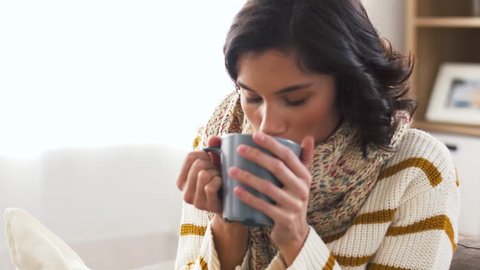 health, cold and people concept - sick young woman in scarf drinking hot tea at home